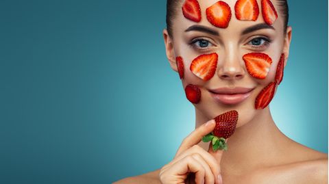 Eine Frau hat Erdbeerscheiben im Gesicht und hält eine Erdbeere in der Hand.
