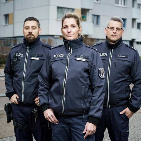 Drei Berliner Polizisten (zwei Männer und eine Frau) stehen vor einem Hochaus in Neukölln