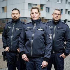 Drei Berliner Polizisten (zwei Männer und eine Frau) stehen vor einem Hochaus in Neukölln