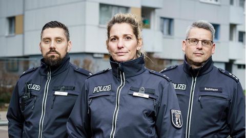 Drei Berliner Polizisten (zwei Männer und eine Frau) stehen vor einem Hochaus in Neukölln