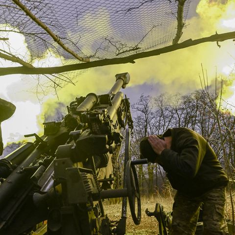 In der Ukraine halten sich zwei Soldaten die Ohren zu, während sie ein Geschütz abfeuern, das einen Feuerball ausstößt