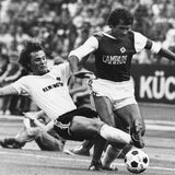 Der Hamburger SV, hier mit Georg "Schorsch Volkert im Einsatz, gehörte ebenfalls zu den Vereinen, die schnell reagierten und ab der Saison 1974/75 die freie Fläche gewinnbringend veräußerten. Für 500.000 DM im Jahr stellten sie ihre Brust für den italienischen Likör Campari zur Verfügung. Der Profi, der hier zu einem Tackling (damals: Grätsche) gegen Volkert ansetzt, ist Bundesliga-Rekordspieler Charlie Körbel von Eintracht Frankfurt. Die SGE gewann als Sponsor den Rasierapparate-Hersteller Remington und gehörte ebenfalls zu den ersten. Vereinen, die die neue Einnahmequelle nutzen.