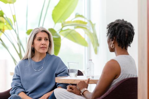 Eine grauhaarige weiße Frau und eine schwarzhaarige schwarze Frau sitzen einander in einer Psychotherapie gegenüber