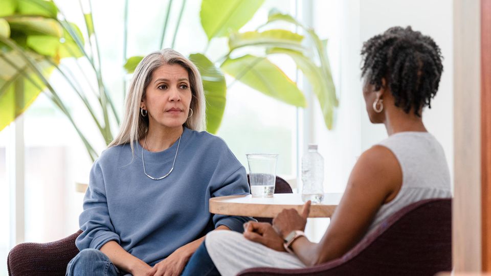 Eine grauhaarige weiße Frau und eine schwarzhaarige schwarze Frau sitzen einander in einer Psychotherapie gegenüber