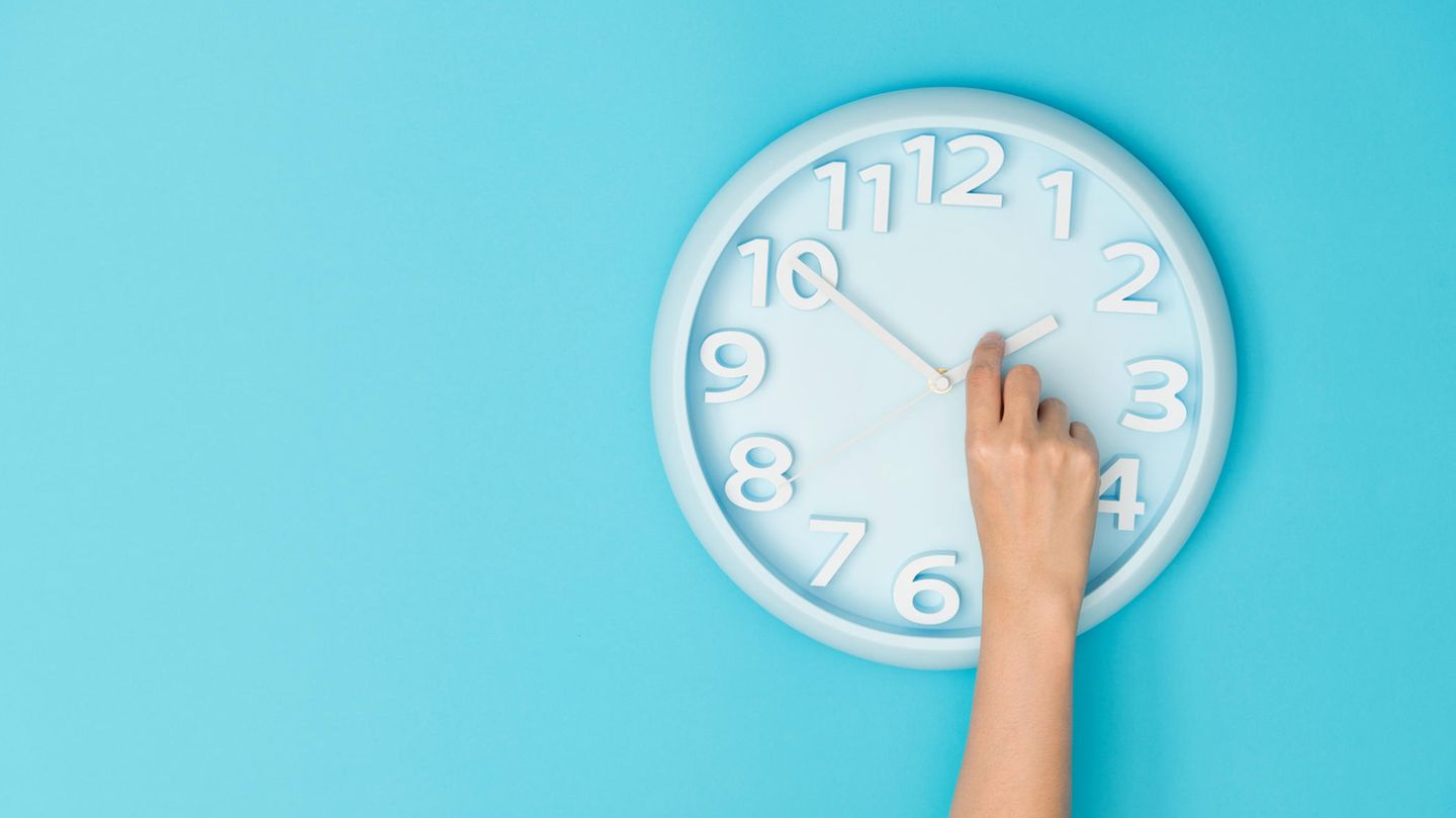 Eine Hand dreht an einer Wanduhr den Stundenzeiger als Symbolfoto für Zeitumstellung