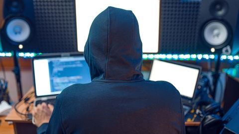 Ein Hacker mit Kapuzenpullover sitzt vor mehreren Laptops an einem Schreibtisch
