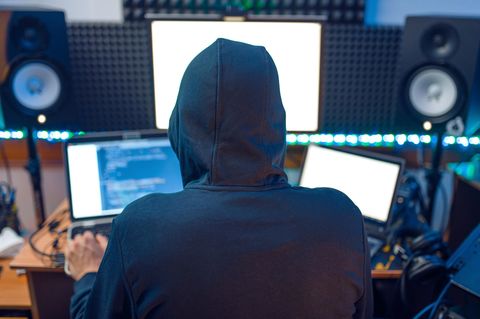 Ein Hacker mit Kapuzenpullover sitzt vor mehreren Laptops an einem Schreibtisch