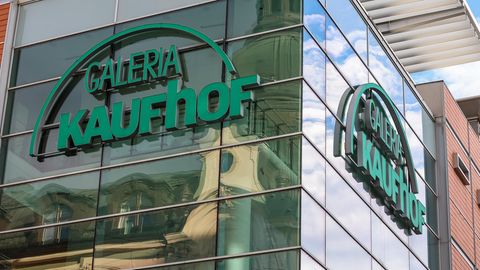 An einer verspiegelten Glasfassade hängt das grüne Logo von Galeria Kaufhof
