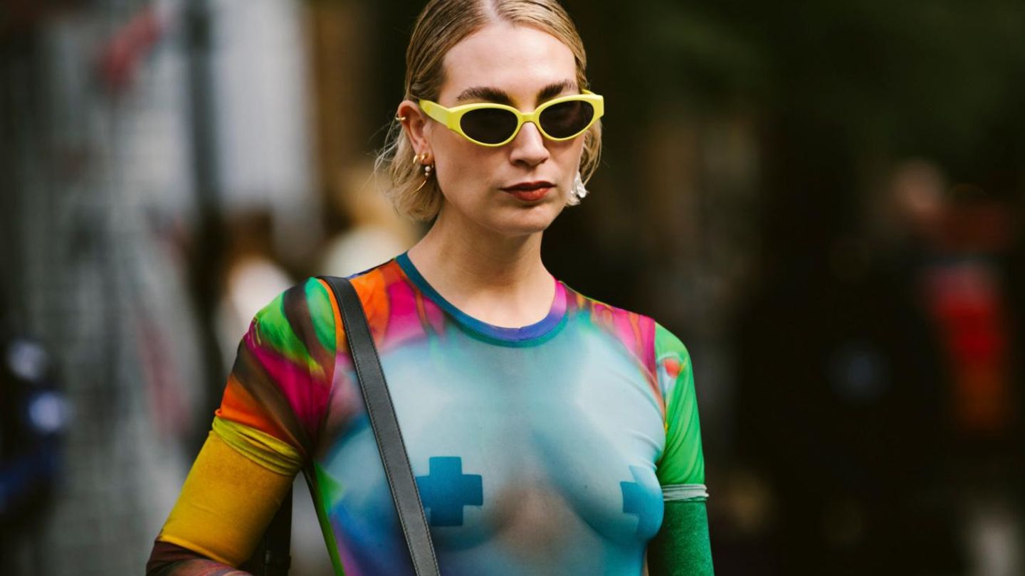 Eine Frau mit einer gelben Sonnenbrille und einem durchsichtigen Netzoberteil mit Nippelpatches