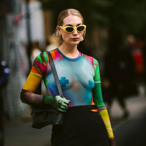 Eine Frau mit einer gelben Sonnenbrille und einem durchsichtigen Netzoberteil mit Nippelpatches