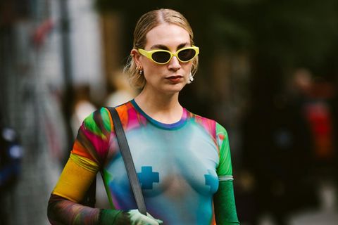 Eine Frau mit einer gelben Sonnenbrille und einem durchsichtigen Netzoberteil mit Nippelpatches