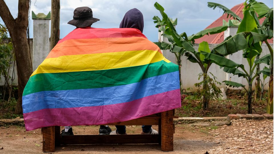 Zwei Personen in Uganda sind von hinten auf einer Bank zu sehen, ihre Rücken bedeckt von einer Regenbogen-Flagge