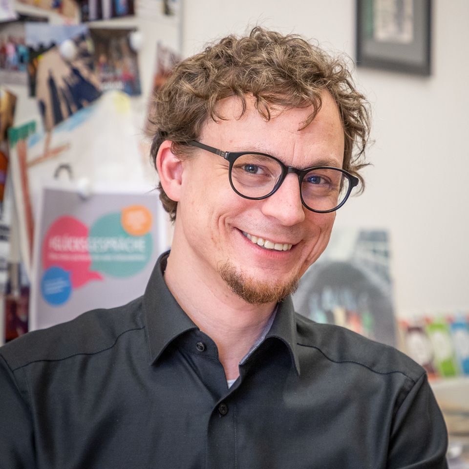 Glücksforscher und Psychologe Tobias Rahm, ein Mann im schwarzen Hemd, mit Brille und Locken, lächelt in die Kamera