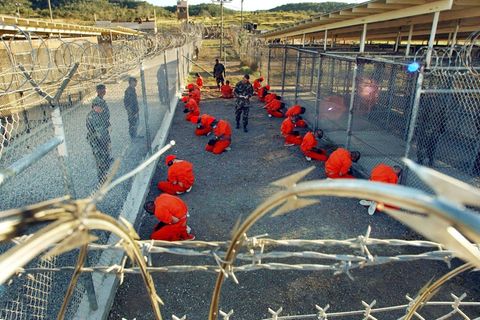 In Guantánamo knien Häftlinge in orange-roten Overalls in einem Verschlag zwischen Drahtzäunen unter freiem Himmel