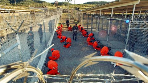 In Guantánamo knien Häftlinge in orange-roten Overalls in einem Verschlag zwischen Drahtzäunen unter freiem Himmel