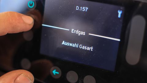 An einer Gasheizung liegen Daumen und Zeigefinger einer linken Männerhand auf einem Display mit Anzeige "Erdgas"