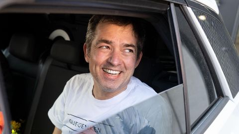 Michael Ballweg sitzt in weißem T-Shirt auf der Rückbank eines weißen PKW und lächelt