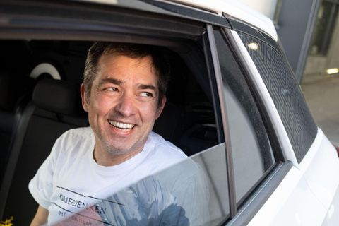 Michael Ballweg sitzt in weißem T-Shirt auf der Rückbank eines weißen PKW und lächelt