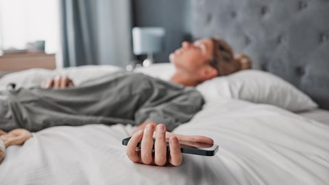 Thema Arbeitssucht: Eine Frau liegt auf dem Bett und hält ihr Handy in der Hand