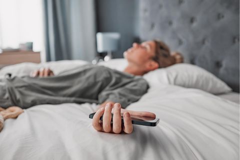 Thema Arbeitssucht: Eine Frau liegt auf dem Bett und hält ihr Handy in der Hand