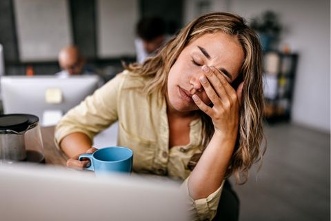 Thema Arbeitssucht: Eine Frau sitzt müde vor ihrem Laptop mit einer Tasse in der Hand