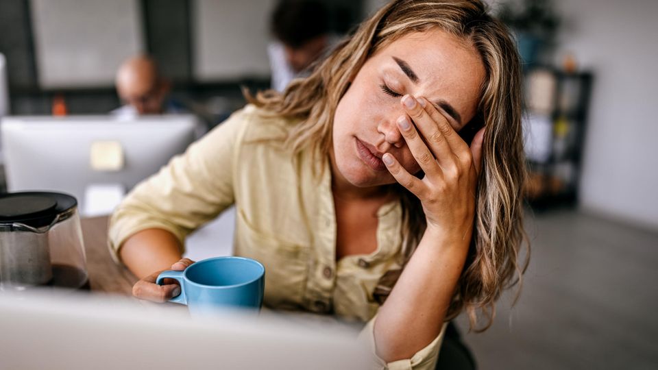 Thema Arbeitssucht: Eine Frau sitzt müde vor ihrem Laptop mit einer Tasse in der Hand