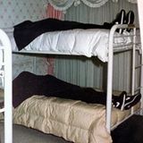 Zwei zugedeckte Tote liegen auf einem Stockbett, sie tragen identische Kleidung und sind mit einem lila Tuch bedeckt