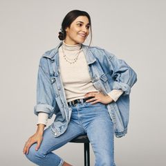 Eine Frau sitzt auf einem Hocker und trägt Jeanshose und -jacke.