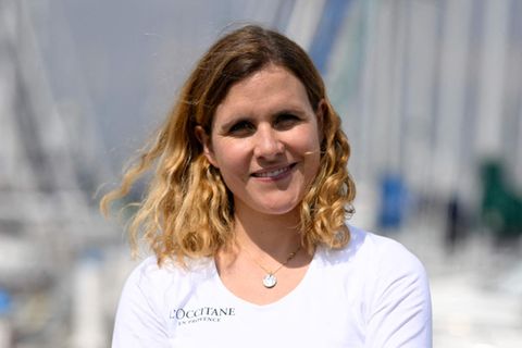 Clarisse Crémer, eine weiße, blonde Frau steht in weißem Langarmshirt in einem Yachthafen und lächelt in die Sonne