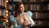 Floridas Gouverneur im sanften blau-goldenen Kleid, tiefen Dekolleté und umgeben von Büchern. Seine Hände sind verschränkt.