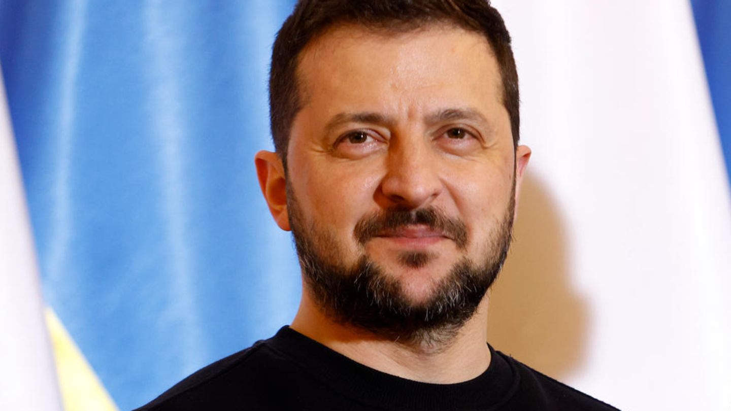 Wolodymyr Selenskyj lächelt leicht, steht vor der Flagge der Ukraine