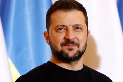Wolodymyr Selenskyj lächelt leicht, steht vor der Flagge der Ukraine