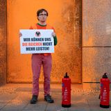 Ein Klimaaktivist der Letzten Generation posiert vor der orangenen Prada-Fassade. Auf einem Schild steht: "Wir können uns die Reichen nicht mehr leisten."