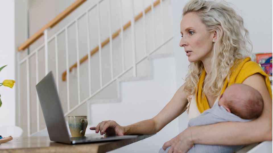 Frau sitzt mit Baby auf dem Arm vor einem Laptop