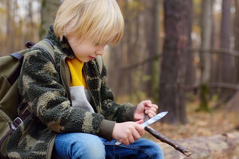 Schnitzmesser für Kinder: Kleiner Pfadfinder sitzt im Wald auf einem Baumstamm und schnitzt an einem Stock