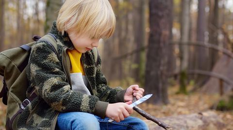 Schnitzmesser für Kinder: Kleiner Pfadfinder sitzt im Wald auf einem Baumstamm und schnitzt an einem Stock