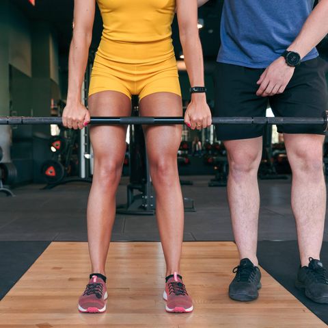 Durchtrainierte Frau beim Krafttraining hält mit gestreckten Armen eine Langhantel, ein Trainer erklärt die Übung
