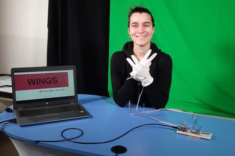Junge Frau sitzt an einem Tisch und hat Handschuhe an die mit einem Laptop verbunden sind