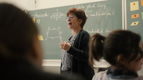 Eine ältere Frau steht vor einer Schulklasse an der Tafel