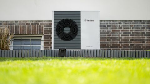 Eine Wärmepumpe - ein weiß-grauer Kasten mit Ventilator - hängt vor dem Backstein-Sockel eines weiß verputzten Wohnhauses