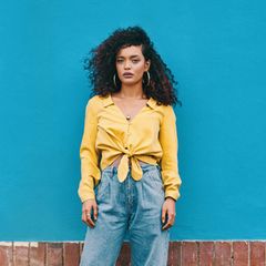 Eine Frag steht vor einem blauen Hintergrund, trägt eine Jeans und eine gelbe Bluse