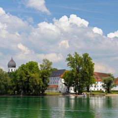 Vorne der See, im Hintergrund das grüne Ufer, ein Kirchturm und weiße Gebäude