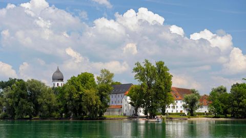 Vorne der See, im Hintergrund das grüne Ufer, ein Kirchturm und weiße Gebäude