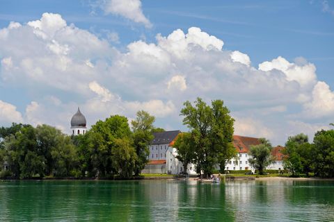 Vorne der See, im Hintergrund das grüne Ufer, ein Kirchturm und weiße Gebäude