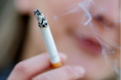 Rauchen: Rauch steigt von der Glut einer Zigarette auf, die eine junge Frau in der rechten Hand hält