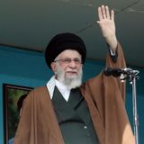Ali Khamenei steht am offenen Fenster vor einem Mikrofon und winkt mit seiner linken Hand. Er trägt einen schwarzen Turban
