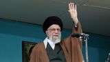 Ali Khamenei steht am offenen Fenster vor einem Mikrofon und winkt mit seiner linken Hand. Er trägt einen schwarzen Turban