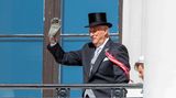 Harald von Norwegen winkt in Frack und Zylinder von einem Balkon, während sein linker Arm sich auf eine Krücke stützt