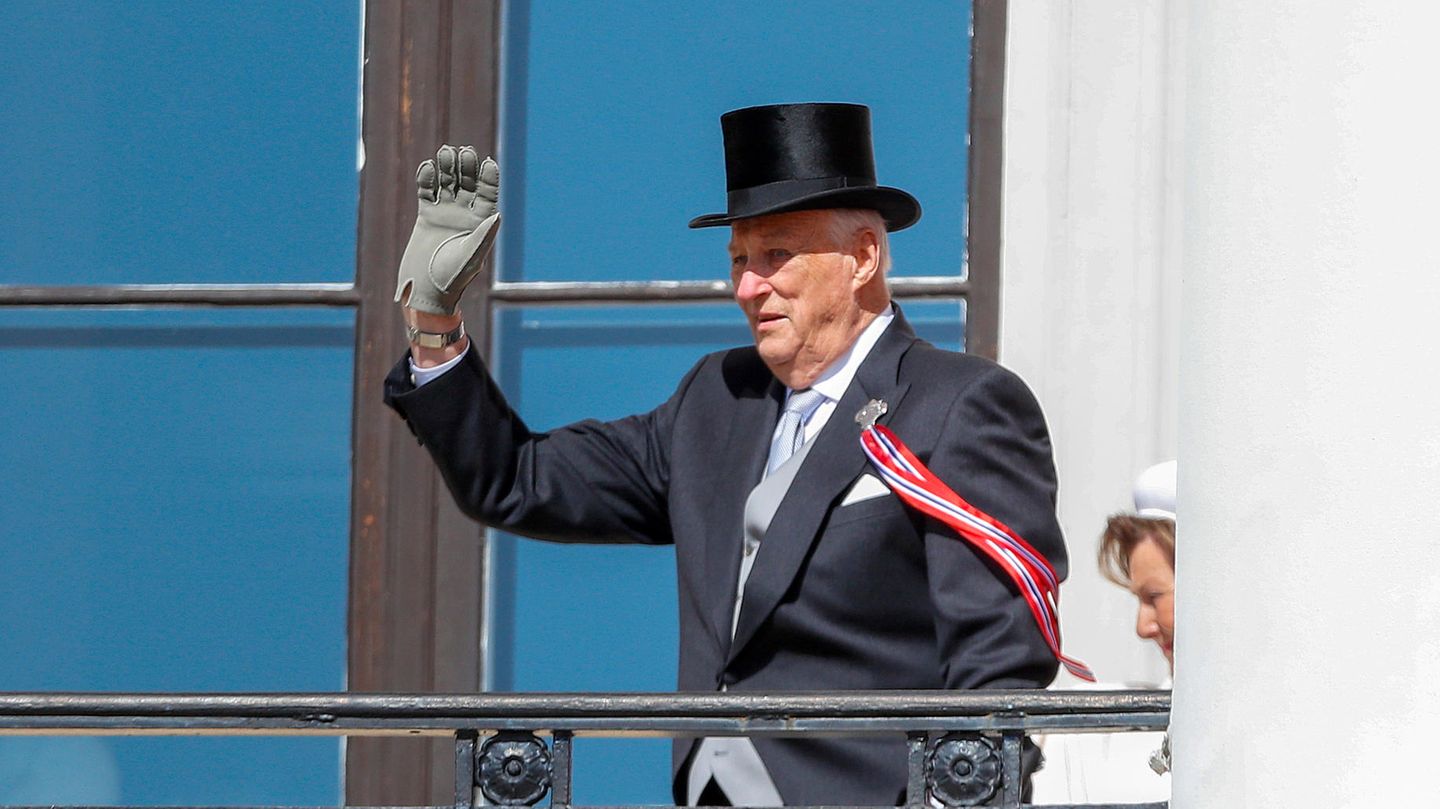 Harald von Norwegen winkt in Frack und Zylinder von einem Balkon, während sein linker Arm sich auf eine Krücke stützt