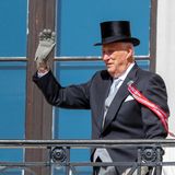 Harald von Norwegen winkt in Frack und Zylinder von einem Balkon, während sein linker Arm sich auf eine Krücke stützt